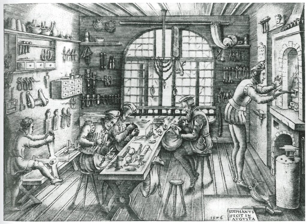 Five people are working inside a workshop making jewelry. One person is working at a forge, while another young person is spinning a wheel of some sort to twist metal. The other three people are working t a long bench.